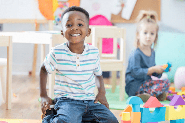Young boy sitting on the floor smiling while another young child plays with blocks in the background