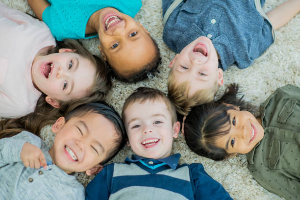 A multi-ethnic group group of young children lay down in a circle on a living room carpet with their heads together all brightly smiling at the camera.