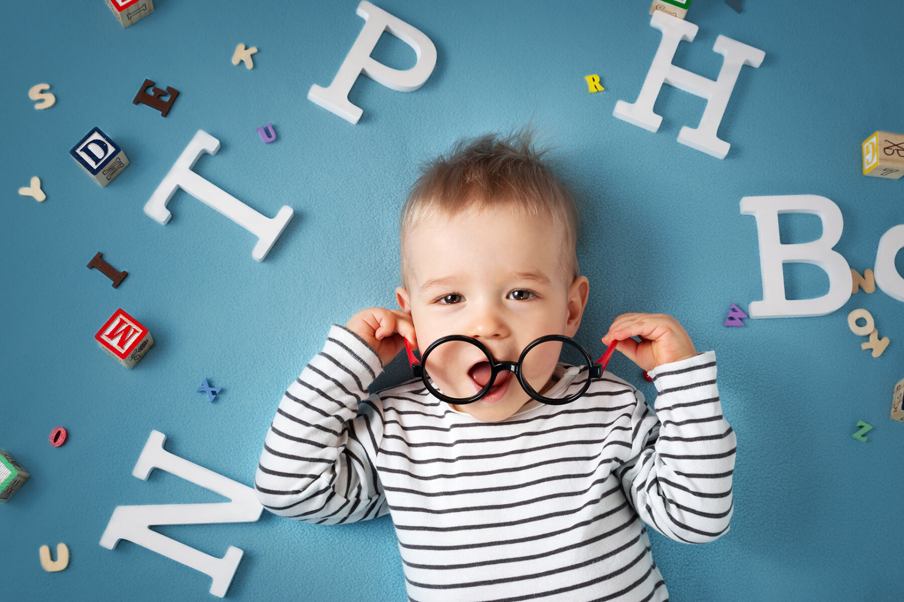 Infant boy looking up while holding glasses surrounded by letters and blocks