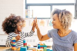 Mother gives her smiling daughter a high five while at the table with building blocks. Parent involvement in ABA therapy.