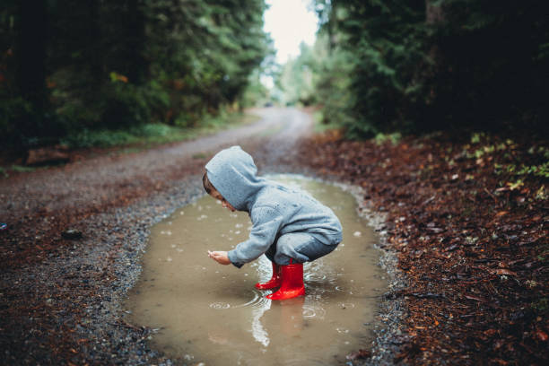 Young boy wearing red rainboots stands in puddle searching for something in the water