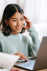 Young woman smiles as she sits scrolling on her computer.