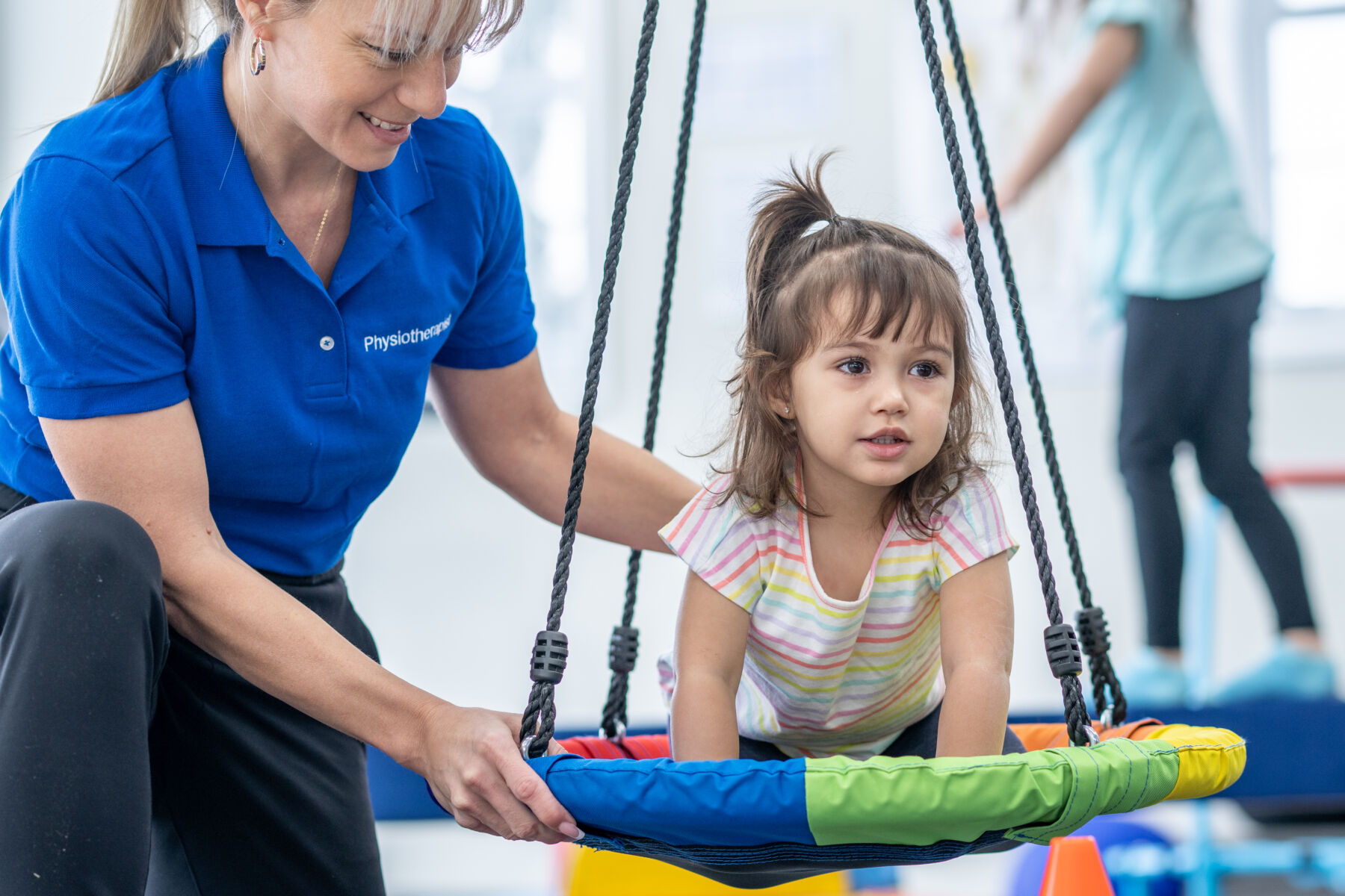 A sweet little girl sits on a swing during a physical therapy session.  She is dressed casually and her female therapist is holding the swing to help steady it as they work on balance together.