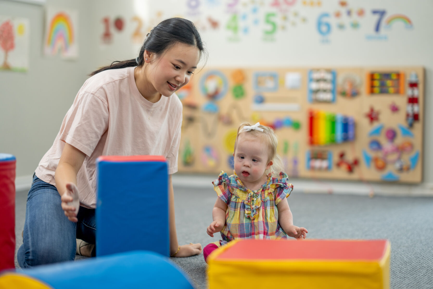 A sweet little girl with Down Syndrome plays on the floor with her Daycare worker, as they work on some gross motor skills with foam blocks.  The little girl is dressed casually and focused on the task.